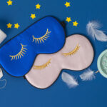 A flat lay of sleep essentials rests against a deep blue background: two silky eye masks embroidered with closed golden lashes, one in midnight blue and the other in soft blush; a vintage mint-green alarm clock frozen at an early hour; scattered white tablets and a teabag suggesting nighttime rituals; delicate white feathers drifting like fragments of dreams; and small yellow stars placed playfully around the scene. The composition evokes the quiet architecture of restful sleep and the circadian cues that guide the body’s nightly restoration.