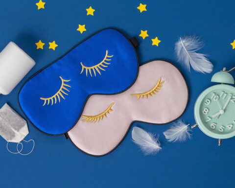 A flat lay of sleep essentials rests against a deep blue background: two silky eye masks embroidered with closed golden lashes, one in midnight blue and the other in soft blush; a vintage mint-green alarm clock frozen at an early hour; scattered white tablets and a teabag suggesting nighttime rituals; delicate white feathers drifting like fragments of dreams; and small yellow stars placed playfully around the scene. The composition evokes the quiet architecture of restful sleep and the circadian cues that guide the body’s nightly restoration.