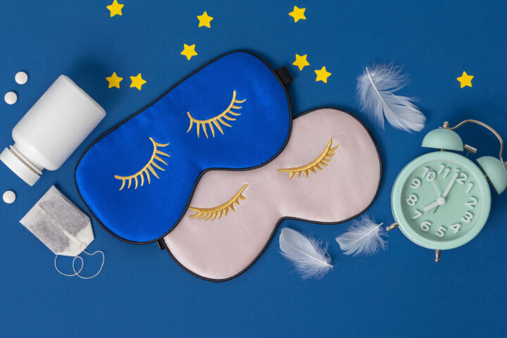 A flat lay of sleep essentials rests against a deep blue background: two silky eye masks embroidered with closed golden lashes, one in midnight blue and the other in soft blush; a vintage mint-green alarm clock frozen at an early hour; scattered white tablets and a teabag suggesting nighttime rituals; delicate white feathers drifting like fragments of dreams; and small yellow stars placed playfully around the scene. The composition evokes the quiet architecture of restful sleep and the circadian cues that guide the body’s nightly restoration.