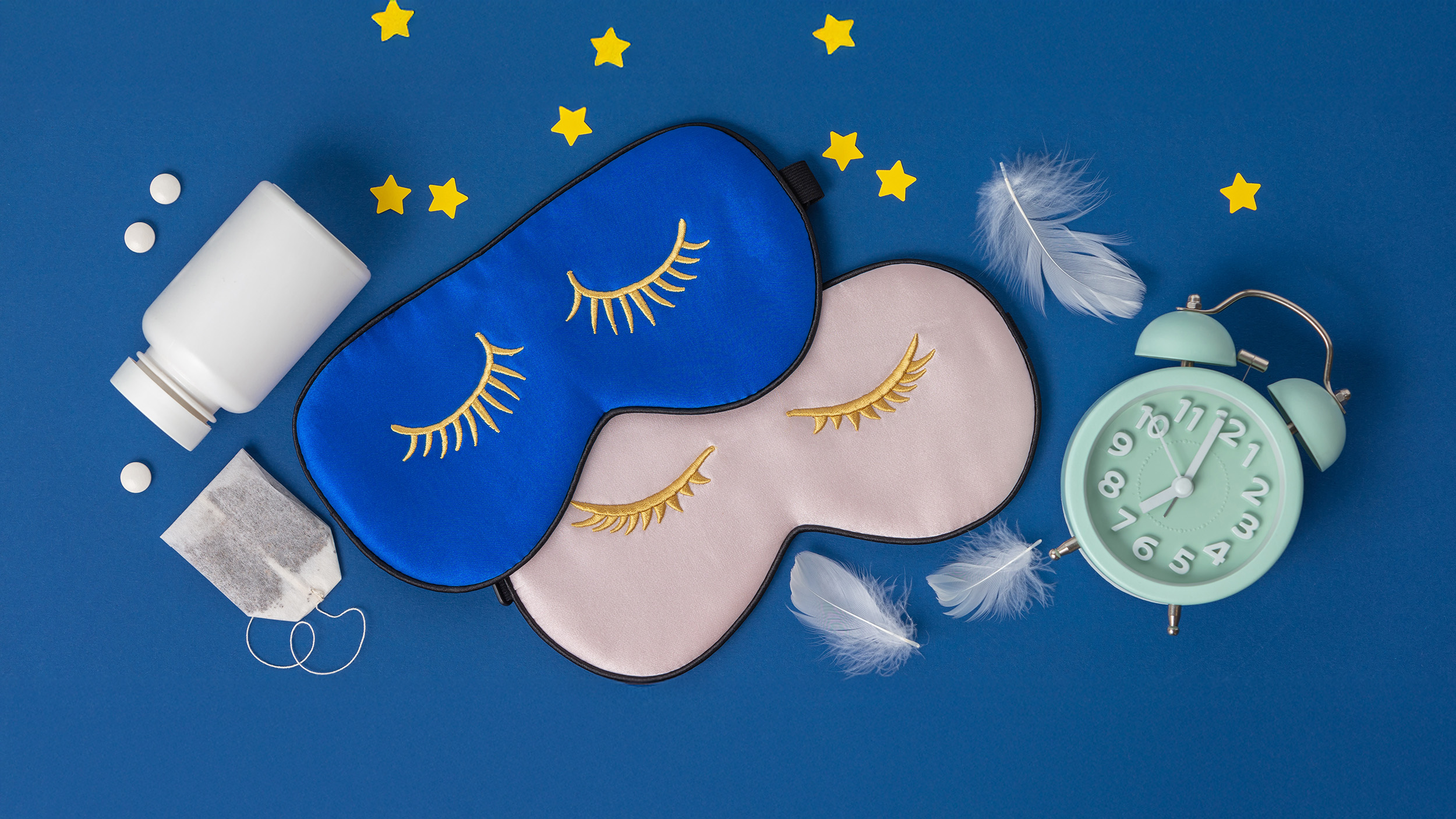 A flat lay of sleep essentials rests against a deep blue background: two silky eye masks embroidered with closed golden lashes, one in midnight blue and the other in soft blush; a vintage mint-green alarm clock frozen at an early hour; scattered white tablets and a teabag suggesting nighttime rituals; delicate white feathers drifting like fragments of dreams; and small yellow stars placed playfully around the scene. The composition evokes the quiet architecture of restful sleep and the circadian cues that guide the body’s nightly restoration.