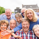 A small circle of older friends gathers outdoors beneath a canopy of warm evening light, faces alive with laughter and recognition. Their hands rest gently on each other’s shoulders as if closing an invisible circuit of care. Behind them, the soft blur of trees and sky seems to echo their calm vitality—a quiet visual proof that belonging itself is a form of medicine. The scene radiates the essence of social embeddedness: the way human connection slows the passage of time and nourishes longevity more deeply than any supplement ever could.