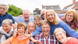 A small circle of older friends gathers outdoors beneath a canopy of warm evening light, faces alive with laughter and recognition. Their hands rest gently on each other’s shoulders as if closing an invisible circuit of care. Behind them, the soft blur of trees and sky seems to echo their calm vitality—a quiet visual proof that belonging itself is a form of medicine. The scene radiates the essence of social embeddedness: the way human connection slows the passage of time and nourishes longevity more deeply than any supplement ever could.