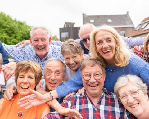 A small circle of older friends gathers outdoors beneath a canopy of warm evening light, faces alive with laughter and recognition. Their hands rest gently on each other’s shoulders as if closing an invisible circuit of care. Behind them, the soft blur of trees and sky seems to echo their calm vitality—a quiet visual proof that belonging itself is a form of medicine. The scene radiates the essence of social embeddedness: the way human connection slows the passage of time and nourishes longevity more deeply than any supplement ever could.
