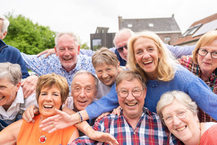 A small circle of older friends gathers outdoors beneath a canopy of warm evening light, faces alive with laughter and recognition. Their hands rest gently on each other’s shoulders as if closing an invisible circuit of care. Behind them, the soft blur of trees and sky seems to echo their calm vitality—a quiet visual proof that belonging itself is a form of medicine. The scene radiates the essence of social embeddedness: the way human connection slows the passage of time and nourishes longevity more deeply than any supplement ever could.