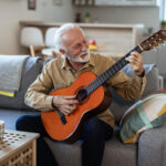 An older man sits on a comfortable living room sofa, gently focused as he practices an acoustic guitar, his posture relaxed but intent. Soft daylight fills the room, and the warm wood of the guitar contrasts with the quiet calm of the space. His expression shows both concentration and joy, capturing the beauty of learning something new later in life — the quiet, meaningful effort that keeps the mind alive and the spirit engaged.