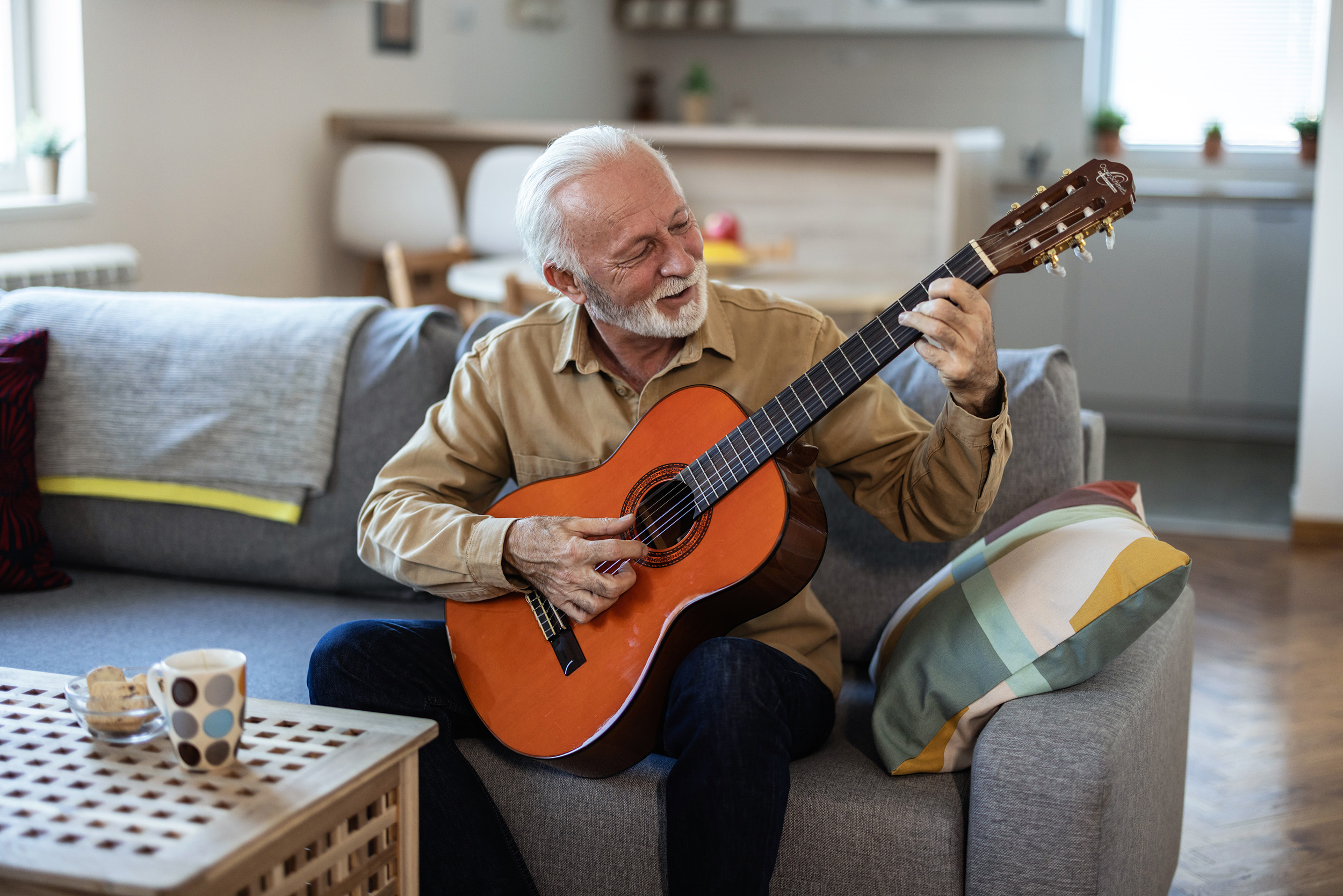 An older man sits on a comfortable living room sofa, gently focused as he practices an acoustic guitar, his posture relaxed but intent. Soft daylight fills the room, and the warm wood of the guitar contrasts with the quiet calm of the space. His expression shows both concentration and joy, capturing the beauty of learning something new later in life — the quiet, meaningful effort that keeps the mind alive and the spirit engaged.