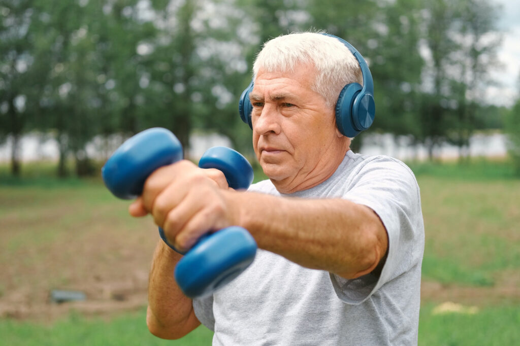 An older man with silver hair exercises outdoors, wearing blue headphones and lifting matching blue dumbbells with focused determination. He stands in a lush green park, his posture strong and steady, embodying resilience, strength, and the ongoing capacity to build muscle and vitality at any age.