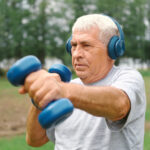 An older man with silver hair exercises outdoors, wearing blue headphones and lifting matching blue dumbbells with focused determination. He stands in a lush green park, his posture strong and steady, embodying resilience, strength, and the ongoing capacity to build muscle and vitality at any age.