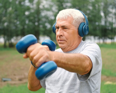 An older man with silver hair exercises outdoors, wearing blue headphones and lifting matching blue dumbbells with focused determination. He stands in a lush green park, his posture strong and steady, embodying resilience, strength, and the ongoing capacity to build muscle and vitality at any age.