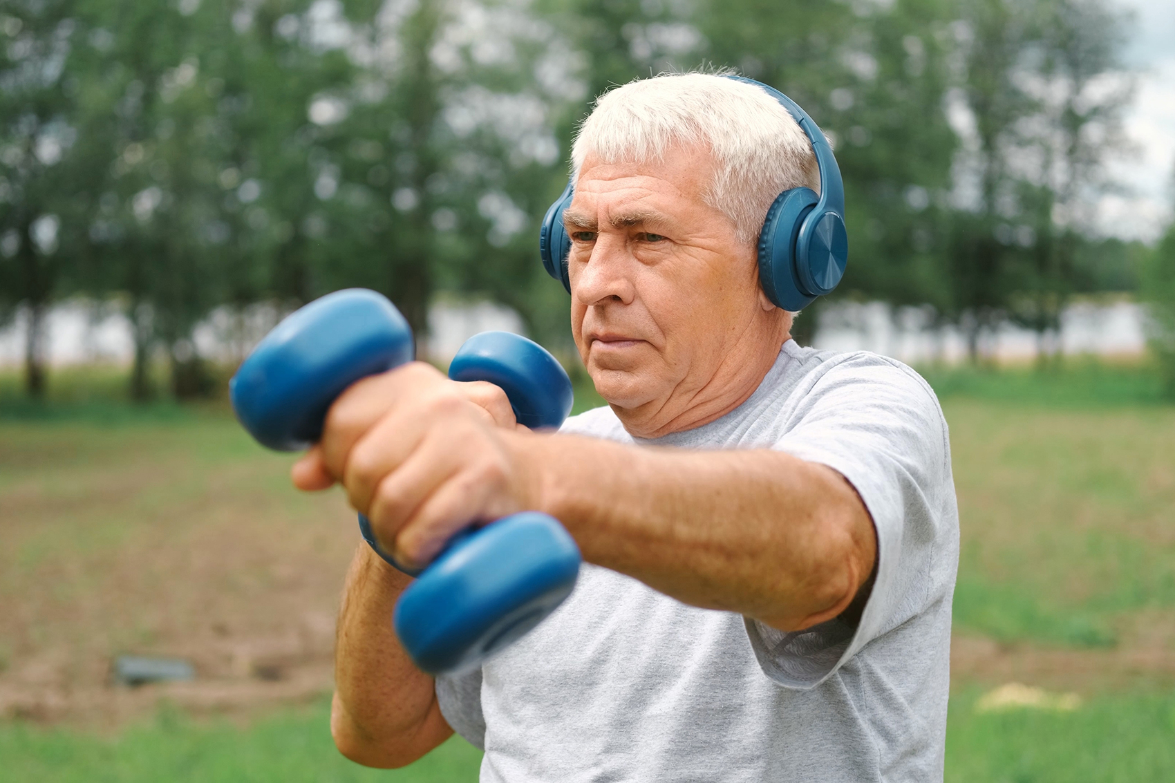 An older man with silver hair exercises outdoors, wearing blue headphones and lifting matching blue dumbbells with focused determination. He stands in a lush green park, his posture strong and steady, embodying resilience, strength, and the ongoing capacity to build muscle and vitality at any age.
