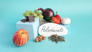 A curated still life of polyphenol-rich foods arranged against a serene turquoise backdrop. A crisp red-striped apple rests in the foreground, while squares of dark chocolate perch on minimalist pale-blue blocks beside hazelnuts, loose green tea leaves, and a handwritten “Polyphenols” label. Behind them, clusters of green grapes, ruby strawberries, a glossy purple onion, and a single white garlic bulb create a vibrant tableau of color and texture, evoking the natural abundance of plant-based compounds that shape human health and longevity.