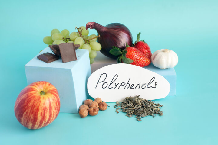 A curated still life of polyphenol-rich foods arranged against a serene turquoise backdrop. A crisp red-striped apple rests in the foreground, while squares of dark chocolate perch on minimalist pale-blue blocks beside hazelnuts, loose green tea leaves, and a handwritten “Polyphenols” label. Behind them, clusters of green grapes, ruby strawberries, a glossy purple onion, and a single white garlic bulb create a vibrant tableau of color and texture, evoking the natural abundance of plant-based compounds that shape human health and longevity.