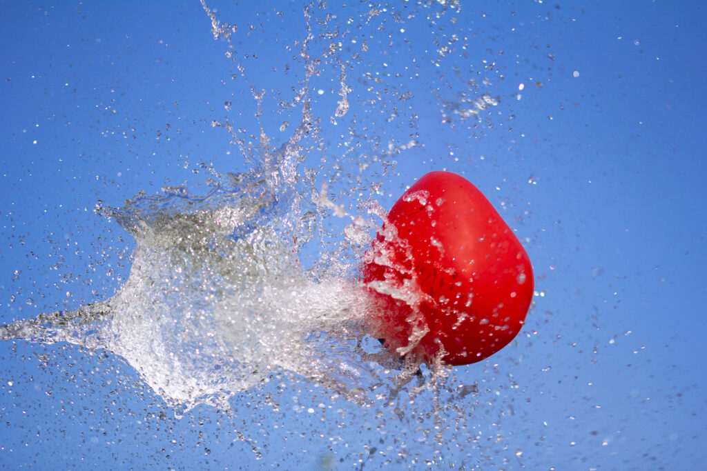 A bright red balloon ruptures midair, frozen in the instant its membrane gives way, sending a crystalline explosion of water outward against a clear blue sky. Droplets scatter in every direction like fractured glass, capturing the drama of a boundary pushed past its limit and the sudden release of what was once contained.