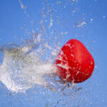 A bright red balloon ruptures midair, frozen in the instant its membrane gives way, sending a crystalline explosion of water outward against a clear blue sky. Droplets scatter in every direction like fractured glass, capturing the drama of a boundary pushed past its limit and the sudden release of what was once contained.