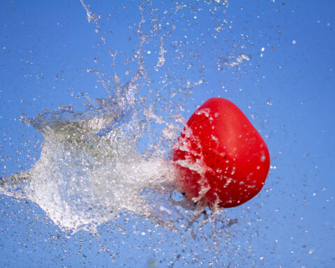 A bright red balloon ruptures midair, frozen in the instant its membrane gives way, sending a crystalline explosion of water outward against a clear blue sky. Droplets scatter in every direction like fractured glass, capturing the drama of a boundary pushed past its limit and the sudden release of what was once contained.